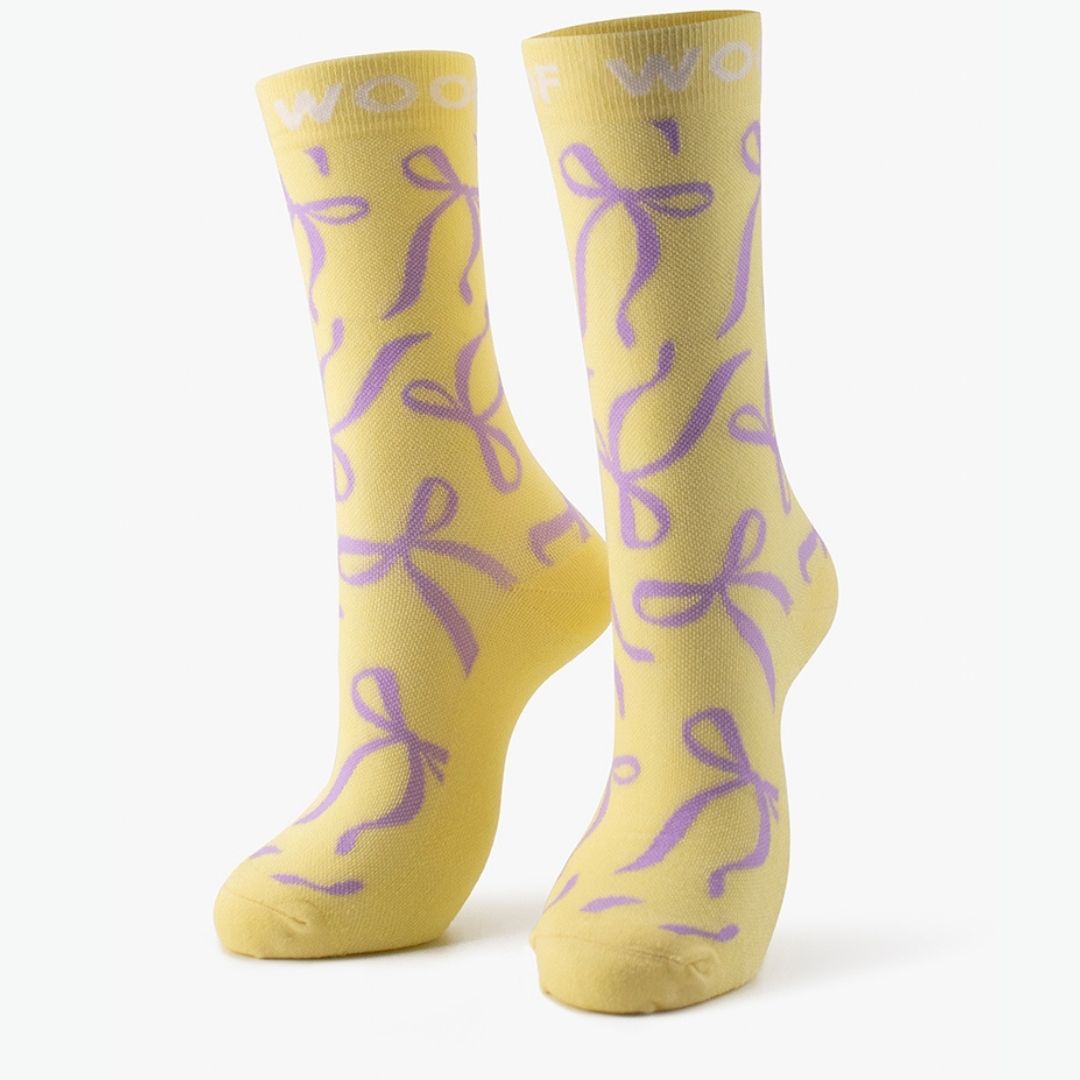 Studio shot of the pair of pastel yellow Bows Bamboo Crew Compression Socks standing upright, featuring purple ribbon bow motifs with white "WOOF" woven into the top cuffs.